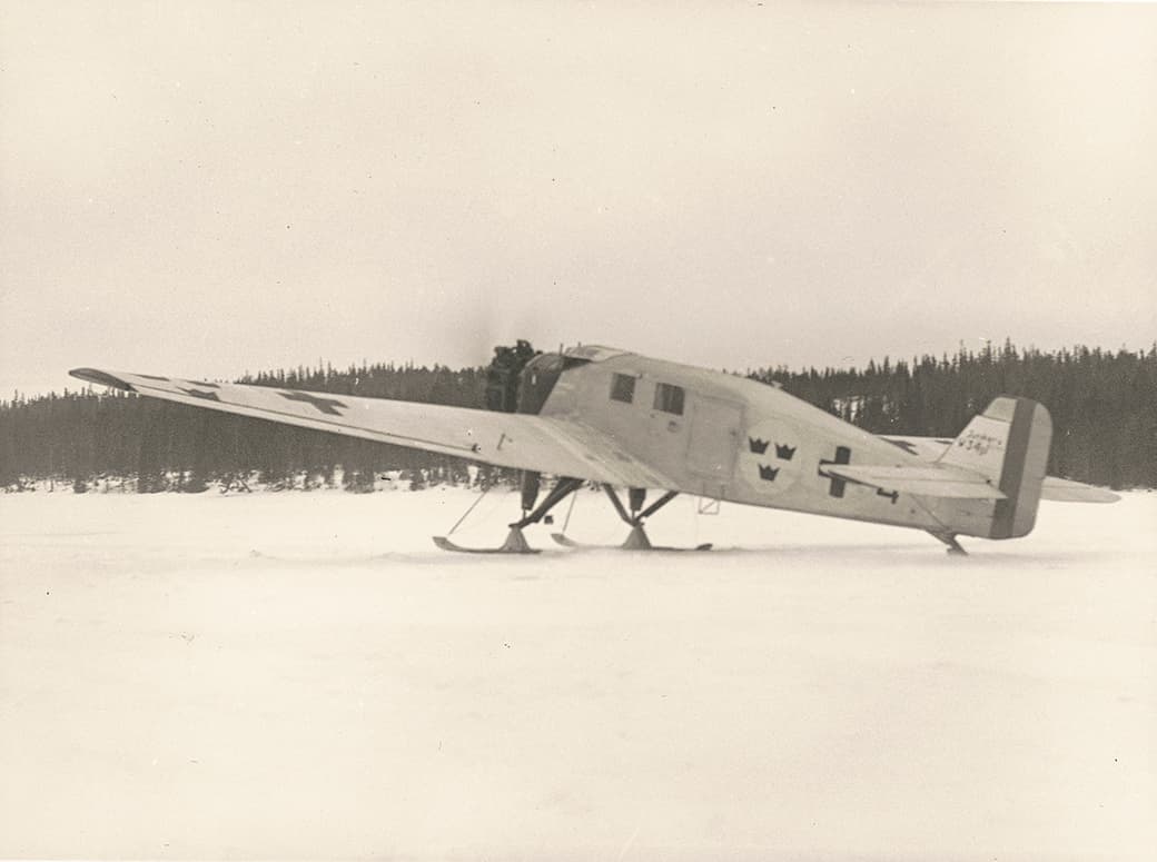 Ett svartvitt fotografi som visar ett äldre ambulansflygplan stående på snötäckt mark, utrustat med skidor för landning. Planet har svenska symboler, inklusive tre kronor, målade på sidan och bakpartiet. En person är synlig vid flygplansdörren, och i bakgrunden skymtar en tät skog. Bilden förmedlar en historisk känsla av tidigt flyg inom vinterlandskap.