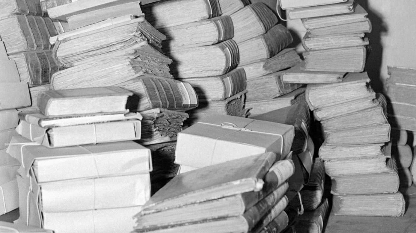 Stora högar med arkivhandlingar och dokumentbuntar. Dokumenten är inbundna och staplade oordnat, vilket skapar en känsla av kaos.