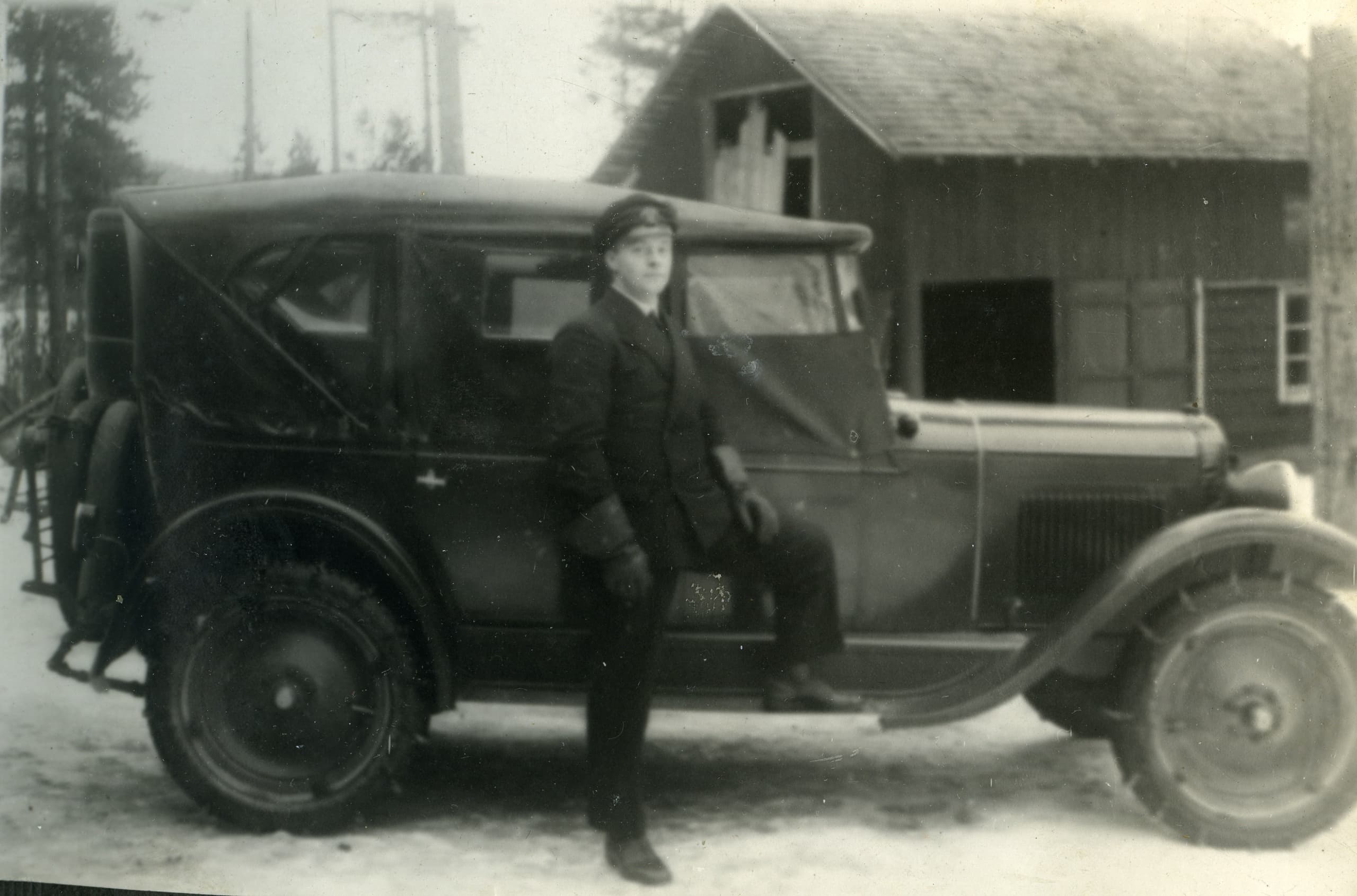 En man i kostym och hatt står framför en bil. Kan vara tidigt 1900-tal.