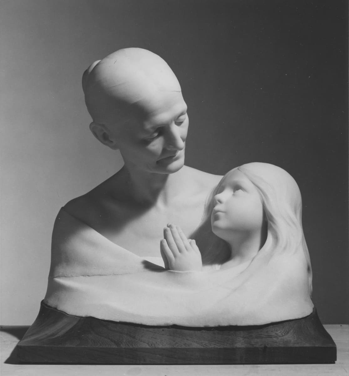 Vit skulptur/byst föreställande ett barn och en vuxen som ser på varandra. Barnet håller ihop sina händer och ser ut att vila i den vuxnes famn.