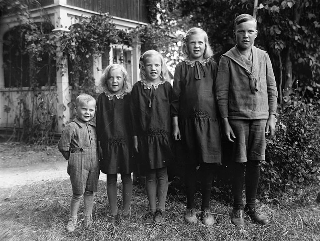 En äldre bild av fem barn i olika åldrar stående utomhus framför ett hus med klättrande växter.