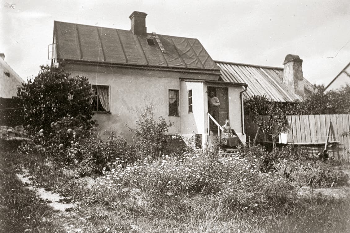 Ett äldre fotografi av ett hus och trädgård med vildvuxna buskar. På trappan till huset sitter en person.