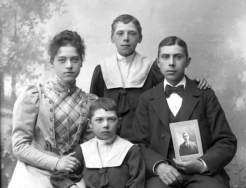 Svartvit porträttbild av en familj från sent 1800-tal eller tidigt 1900-tal, med en kvinna, två unga pojkar och en man som håller ett fotografi i handen.