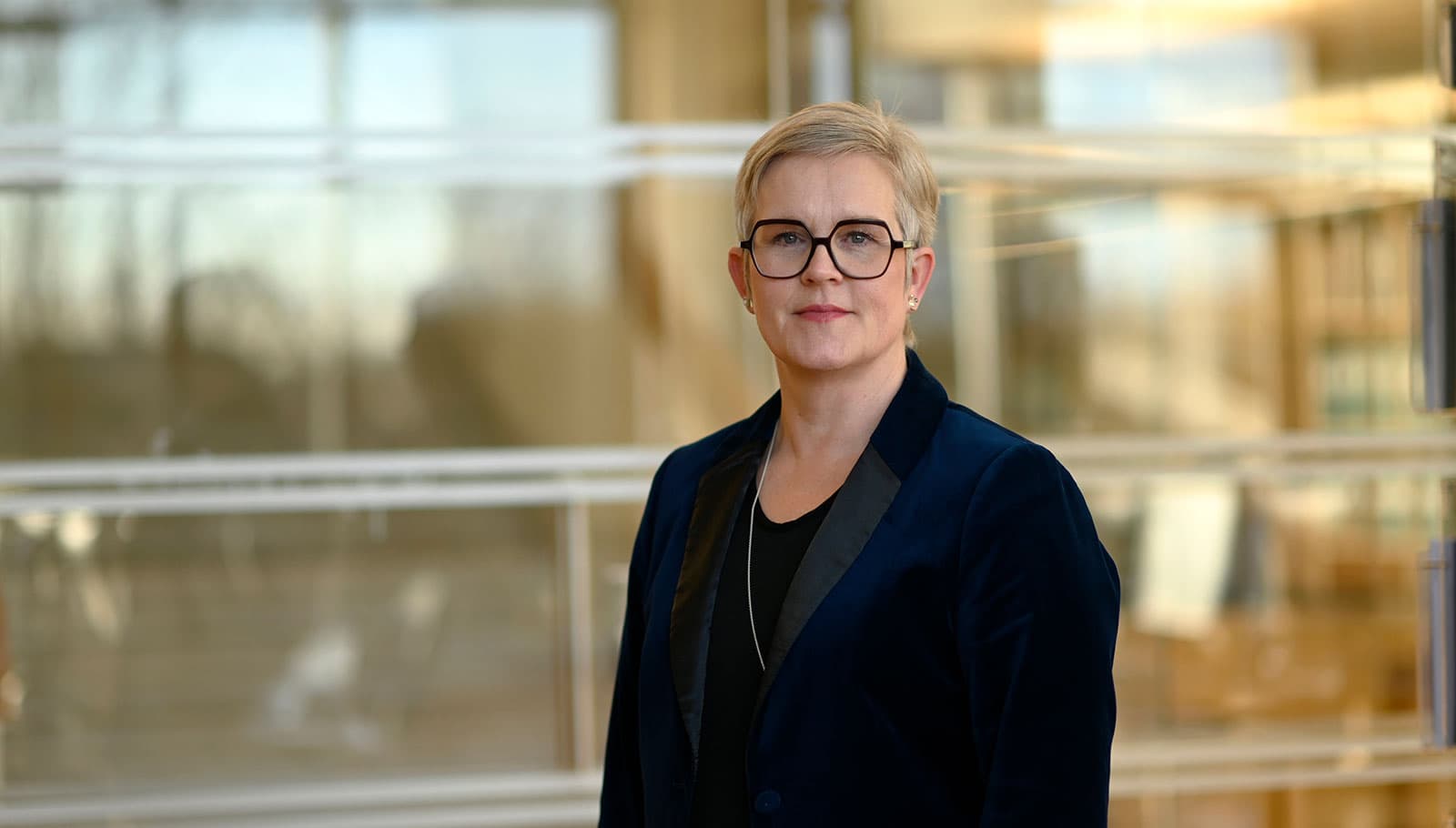 Porträtt av en kvinna med kort blont hår och glasögon, klädd i en mörk kavaj, som står framför en oskarp bakgrund med glaspartier. Hon ser direkt in i kameran med ett neutralt ansiktsuttryck.