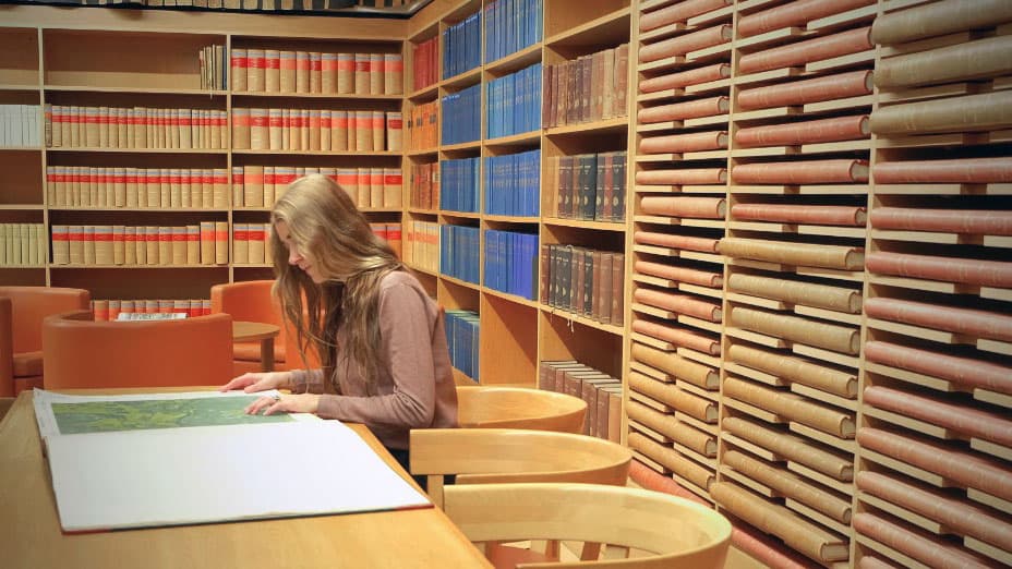 En person sitter vid ett bord och läser i en bok. Längs väggarna finns hyllor emd böcker.